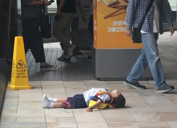 身穿校服的年幼男童在沙田中心對開的行人通道上,背著書包呈大字型橫卧。