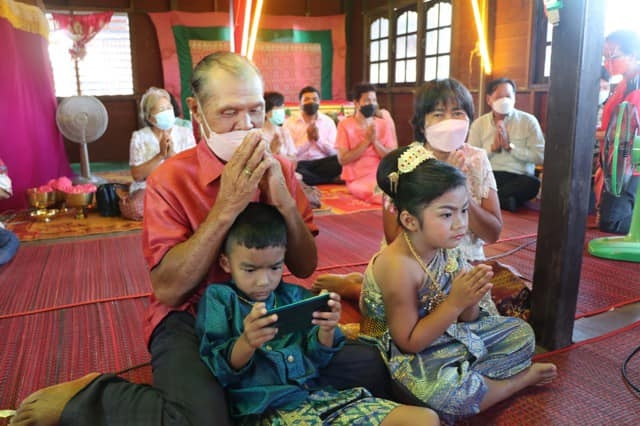 農穆躺在爺爺身上玩手機，農邁雙手合十聽從長輩的指示。